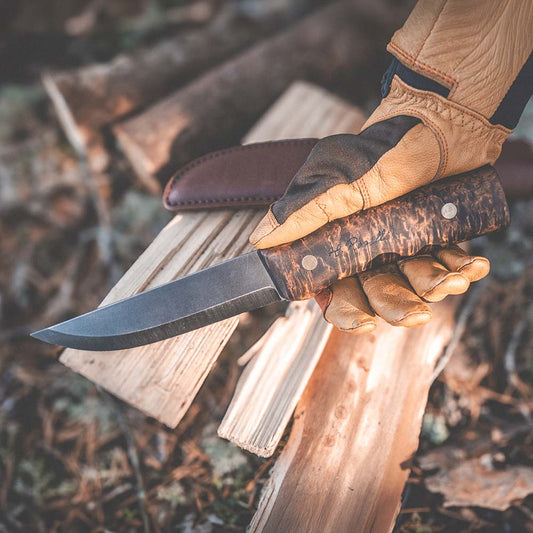A close-up of a hand wearing a tan leather work glove holding a Roselli Hunting Knife Long full tang knife with a dark curly birch handle, positioned over stacked firewood outdoors.