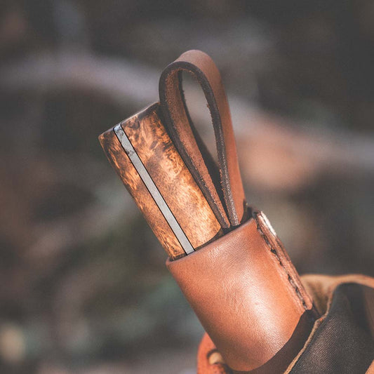 A close-up of a Roselli leather sheath holding a knife with a curly birch handle, set against a blurred outdoor background.